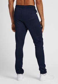 Donkerblauwe joggerbroek met elastische tailleband, taps toelopend ontwerp en zijzak met rits. Witte sneakers gedragen erbij. Subtiele merkdetail zichtbaar.