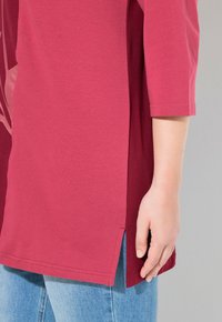 Bordeauxfarbene Tunika mit rundem Halsausschnitt, seitlichem Schlitz und glatter Textur. Kombiniert mit hellblauen Jeans.