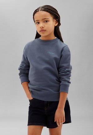 Mädchen mit geflochtenem Haar, das einen blauen Calvin Klein Sweatshirt und schwarze Shorts trägt, steht vor einem schlichten grauen Hintergrund.
