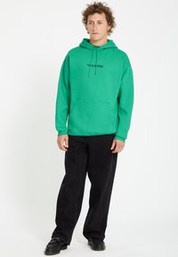 Groene katoenen hoodie met een voorzak, trekkoordcapuchon en zwarte logo-tekst. Gemaakt in combinatie met zwarte broek en zwarte schoenen.