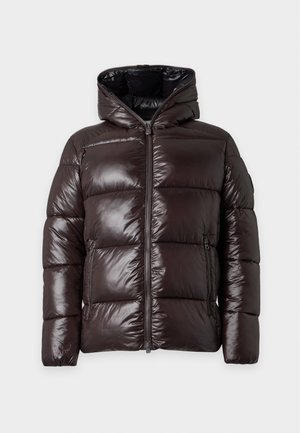 Homme portant une doudoune noire brillante avec capuche, chemise noire, pantalons noirs amples et chaussures en cuir marron, debout sur un fond uni.