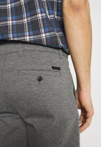 Pantalons gris texturé avec une poche arrière comportant un bouton noir et une petite étiquette de logo. Complète un design de chemise à carreaux.