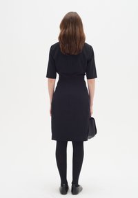 Svart klänning med korta ärmar, figursydd bodice och knälång kjol. Bärs med svarta tights och skor, och håller en svart handväska.