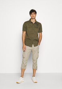 Chemise olive à manches courtes avec deux poches avant et logo noir, associée à un pantalon cargo beige clair, et des baskets blanches avec semelles en caoutchouc.