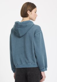 Blauwe, oversized hoodie van een textuurstof, met een verstelbare kap en geribbelde manchetten, gecombineerd met donkere jeans. Zicht van achteren.