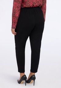 Pantalones negros ajustados con cintura elástica. El modelo lleva zapatos de tacón negros. El fondo es blanco liso.