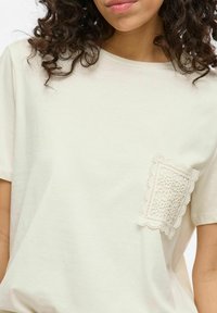 Camiseta de algodón color beige claro con cuello redondo, mangas cortas y un detalle de bolsillo de encaje texturizado en el lado izquierdo del pecho.