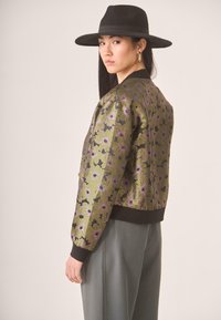 Mujer joven con un sombrero negro de ala ancha, pendientes de aro dorados, chaqueta verde con estampado floral y pantalones grises, mirando por encima del hombro.