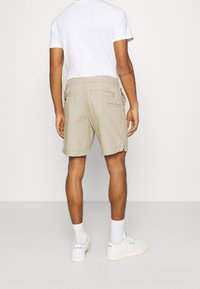 Man wearing beige shorts, white t-shirt, white socks, and white Reebok sneakers, standing facing a plain light-colored wall.