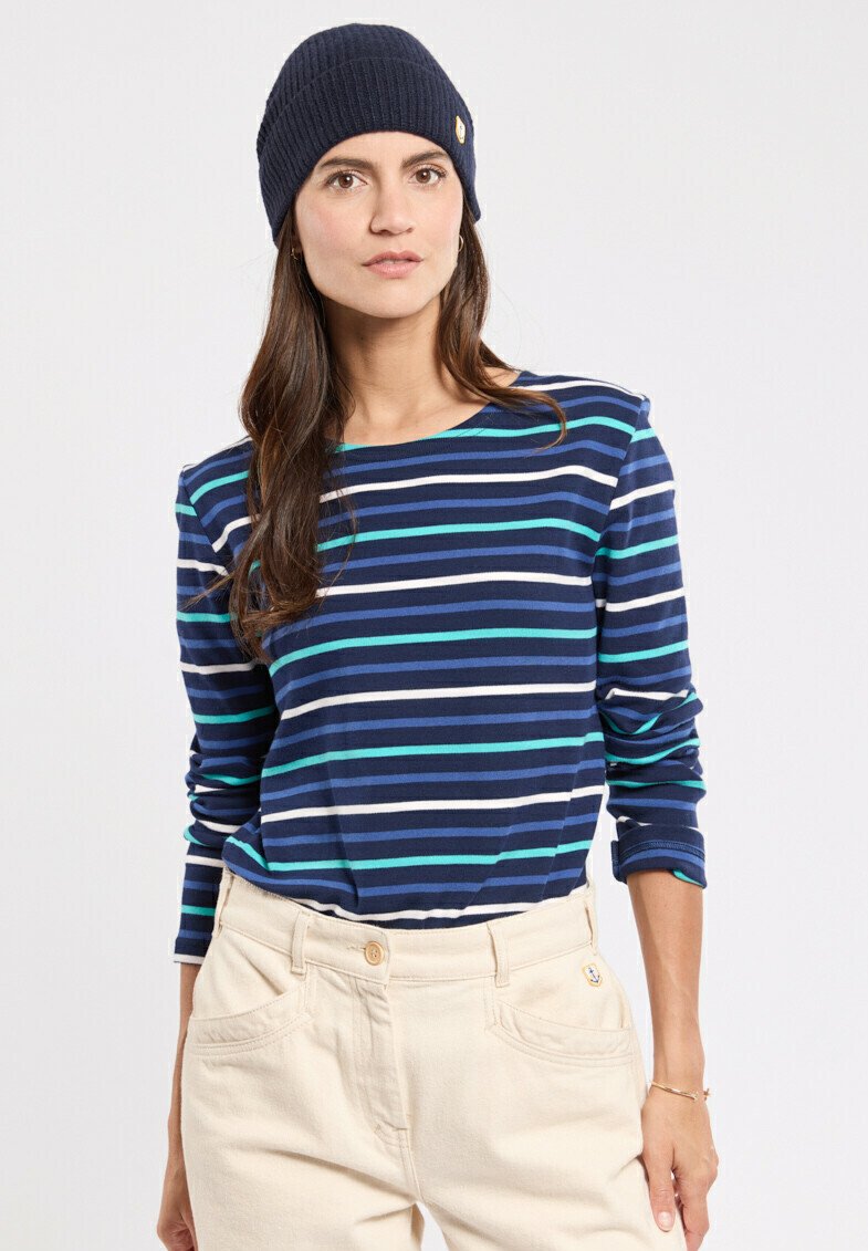 Marineblauw en witte gestreepte langemouwen shirt met turkooisaccenten, gecombineerd met crèmekleurige broek. Het model draagt een marineblauwe ribgebreide beanie.
