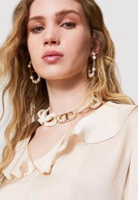 Blusa beige con colletto arricciato, abbinata a una collana di perle e catena dorata. La modella ha i capelli biondi ondulati e orecchini a goccia di perle.
