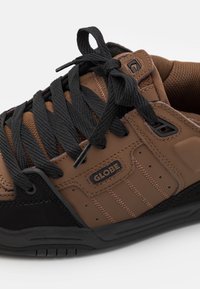 Baskets marron et noires avec cuir texturé et détails en mesh. Elles comportent des lacets noirs épais, un patch logo et un embout renforcé.