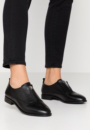 Chaussures en cuir noir avec un bout arrondi et un détail de fermeture éclair sur le côté. Semelle plate, texture lisse et accents de couture minimalistes. Associées à un jean noir.