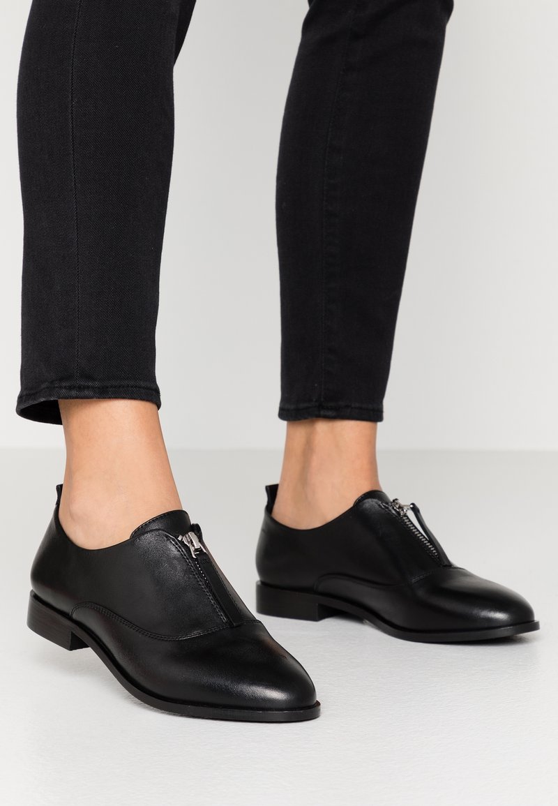 Chaussures en cuir noir avec un bout arrondi et un détail de fermeture éclair sur le côté. Semelle plate, texture lisse et accents de couture minimalistes. Associées à un jean noir.