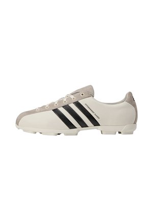 Scarpetta da calcio bianca con strisce nere, dettagli beige in nappa e lacci bianchi, mostrata di profilo destro su sfondo bianco.