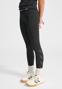 Schwarze athletische Leggings mit hohem Bund und einem strukturierten Chevron-Muster entlang der Außenseite des Beins. Kombiniert mit weißen Turnschuhen.