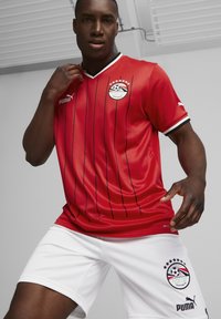 Raudona ir juoda juostuota futbolo marškinėliai su balta apykakle, su emblema ant krūtinės, derinami su baltais šortais su logotipo akcentais.