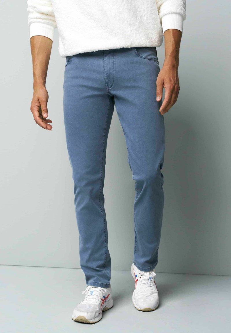 Jean slim bleu clair en denim, avec des poches latérales et une texture lisse, associés à des baskets blanches avec des accents rouges.