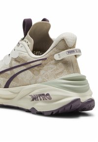 Zapatilla de running beige con parte superior de malla texturizada, acentos morados y una gruesa suela media blanca. Presenta un prominente logo "NITRO" en el talón.