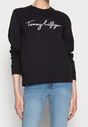 Personne portant un sweat-shirt noir avec un texte blanc "Tommy Hilfiger" et un jean bleu clair sur un fond clair uni.
