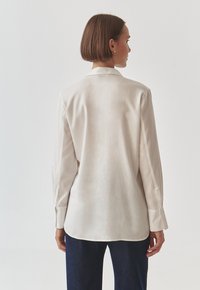 Světle béžová košile s dlouhým rukávem z hladkého materiálu, s klasickým límcem, zapínáním na knoflíky na manžetách a zakřiveným lemem, nošená přes tmavé džíny.