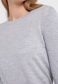 Vaaleanharmaa neulepaita, jossa on pyöreä pääntie, ja joka sisältää sileän pinnan sekä ribbireunukset hihoissa. Lähikuva näyttää yksityiskohtia.