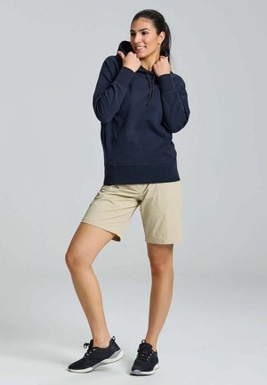 Haut à capuche navy avec poche kangourou, cordon de serrage ajustable, associé à un short beige et des chaussures de sport noires avec des semelles blanches.