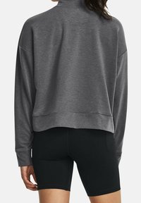 Vrouw in een losse grijze lange mouw top en strakke zwarte fietsshort, gezien vanuit de achterkant tegen een effen achtergrond.