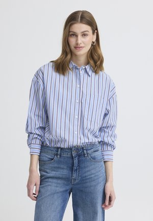 Kvinde med skulderlangt brunt hår iført en blå og hvid lodretstribet skjorte og blå jeans, stående foran en simpel baggrund.