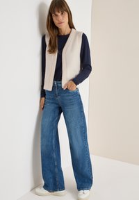 Helles beiges gestricktes ärmelloses Vest, kombiniert mit einem marineblauen Langarmshirt und weiten blauen Jeans. Weiße Sneakers vervollständigen das Outfit.