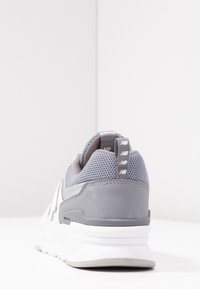 Grå och vit New Balance-sko sedd bakifrån, med fokus på den texturerade textilen, dragfliken med logo och den dämpade sulan.