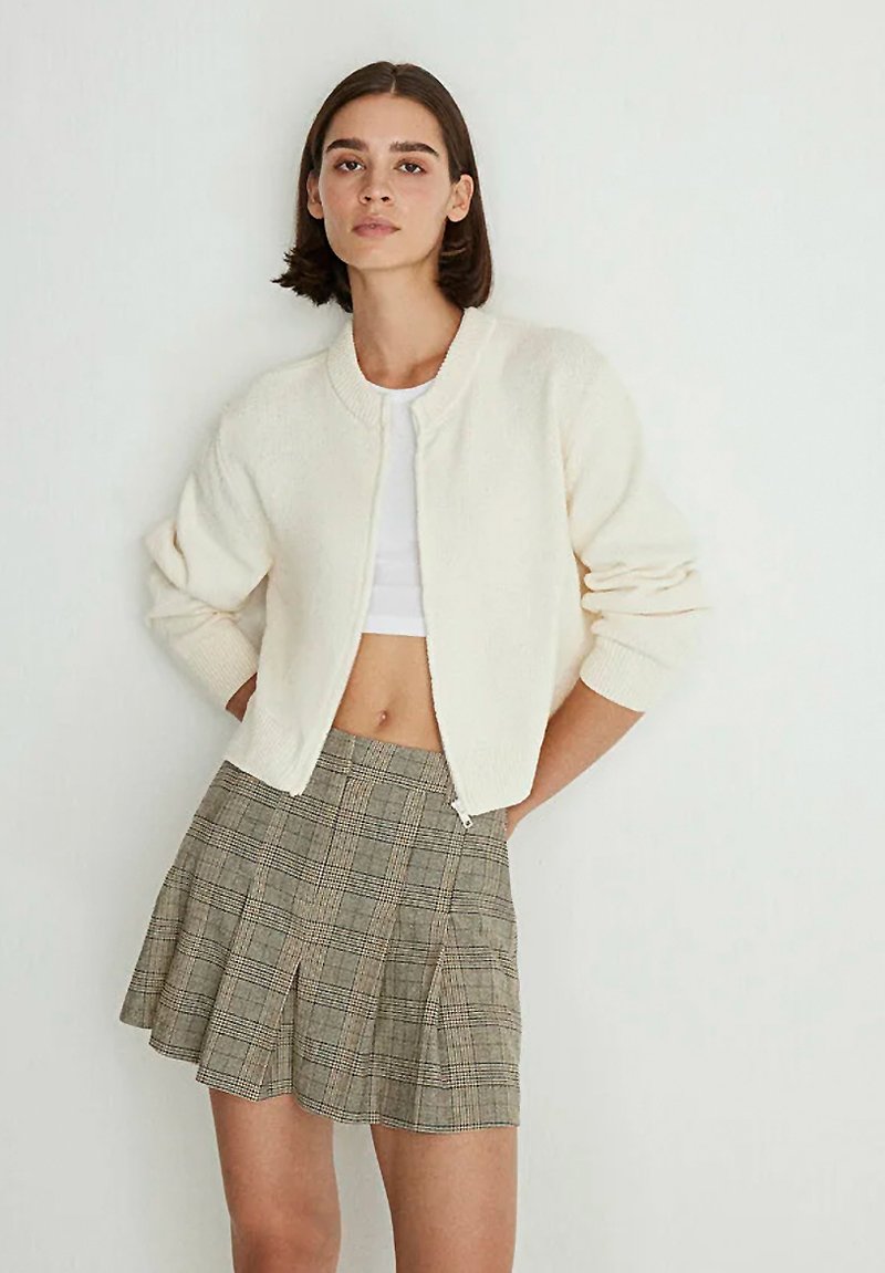 Cream, textured cardigan with a round neck and front zip, paired with a plaid mini skirt featuring pleats and a neutral color palette.