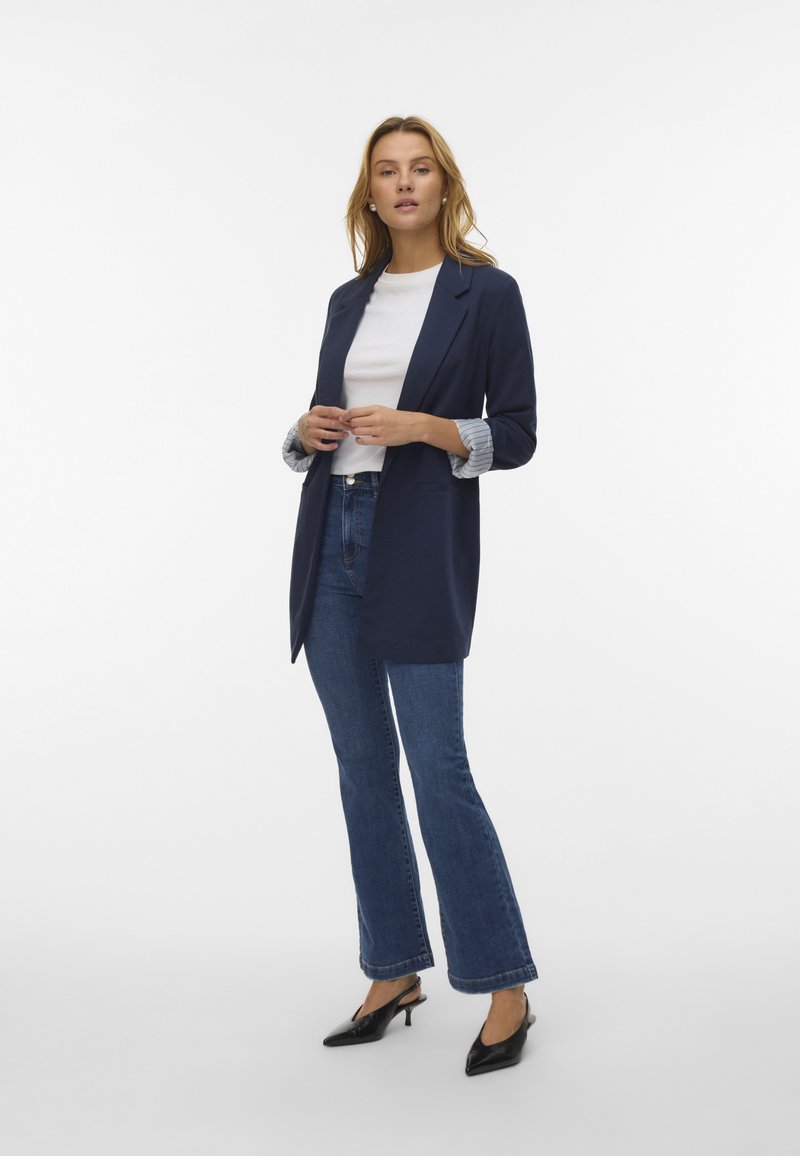 Mornarsko modra blazerjakna z zapiranjem na en gumb, kombinirana z belim zgornjim delom in razširjenimi kavbojkami. Črne čevlje s koničastim nosom zaokrožajo videz.