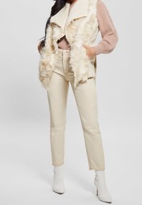 Donna che indossa pantaloni color crema, stivaletti bianchi con tacco, un maglione rosa a coste e un gilet color crema con bordo di pelliccia, con le mani nelle tasche.