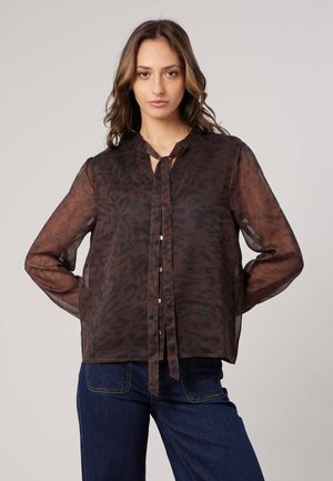 Vrouw die een doorzichtige bruine blouse met patroon draagt met lange mouwen en een strik aan de voorkant, gecombineerd met een blauwe spijkerbroek, staand tegen een effen achtergrond.