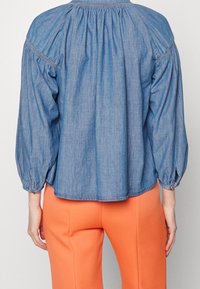 Blusa di denim con schiena arricciata, maniche lunghe a sbuffo e dettagli cuciti. Abbinata a pantaloni arancioni vivaci, creando un look casual.