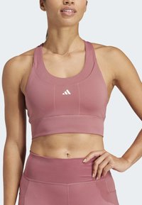 Mujer vestida con un sujetador deportivo de color rosa polvoriento y leggings de cintura alta a juego, con la mano en la cadera contra un fondo sencillo.