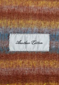 Tekstuuriga puuvillane kangas, millel on rooste, kollase ja sinise värvi gradient, ning valge silt, millel mustade detailidega on õmmeldud "Another Cotton."