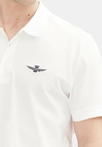 Polo bianco realizzato in tessuto testurizzato, caratterizzato da una grafica ricamata in rilievo di un emblema alato sul lato sinistro del petto.