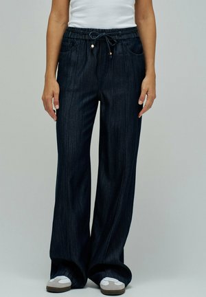 Mujer vestida con jeans oscuros de pierna ancha con cintura elástica y zapatillas blancas, de pie contra un fondo liso.