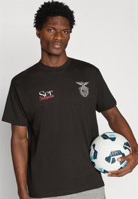 T-shirt de algodão preto com logotipo e texto impressos em branco no peito. O modelo segura uma bola de futebol estampada. Design simples de decote redondo.