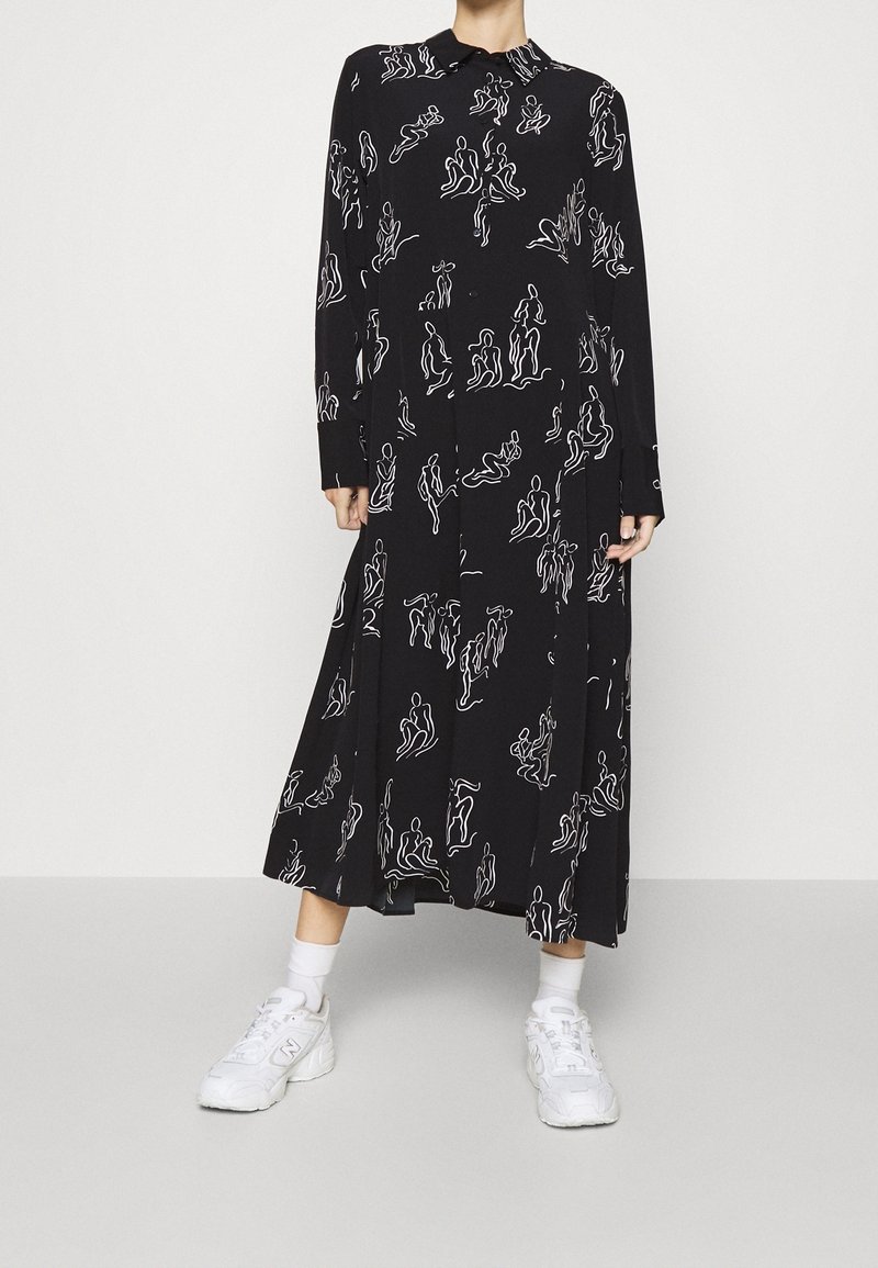 Robe midi noire à manches longues avec des dessins abstraits blancs représentant des silhouettes humaines, portée avec des chaussettes blanches et des baskets blanches sur un fond uni.