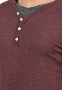 Burgundiszínű henley ing, puha, textúrált anyagból készült, V-nyakkivágással, három fehér gombbal és egy réteges szürke belső inggel.