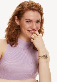 Gouden horloge met een ronde wijzerplaat en schakelarmband, paarse geribbelde top, gelaagd gouden kettingen met kleine accenten en een eenvoudige ring aan de hand.