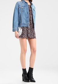 Kvinna iförd en blommig klänning, blå jeansjacka, svarta ankelstövlar och håller en liten silverfärgad rund väska.