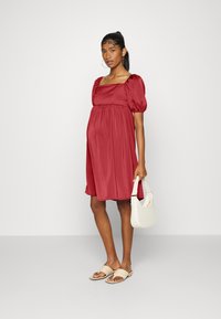 Vestido de seda vermelha com mangas bufantes, decote quadrado e cintura franzida. Modelo segura uma bolsa bege clara e usa sandálias beges.