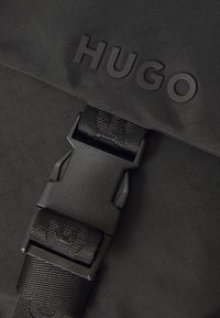 Borsa nera con finitura opaca, logo "HUGO" in rilievo e una robusta tracolla nera con dettagli testurizzati.