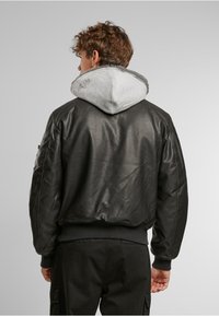Veste aviateur en cuir noir avec une capuche de style sweat-shirt gris et des poignets côtelés, présentant une texture lisse et un design minimaliste.