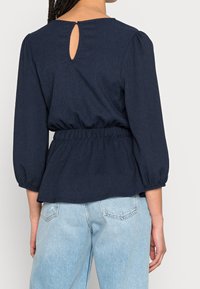 Top peplum blu navy con maniche a tre quarti a palloncino, vita arricciata e dettaglio a chiave sul retro. Tessuto a texture liscia.