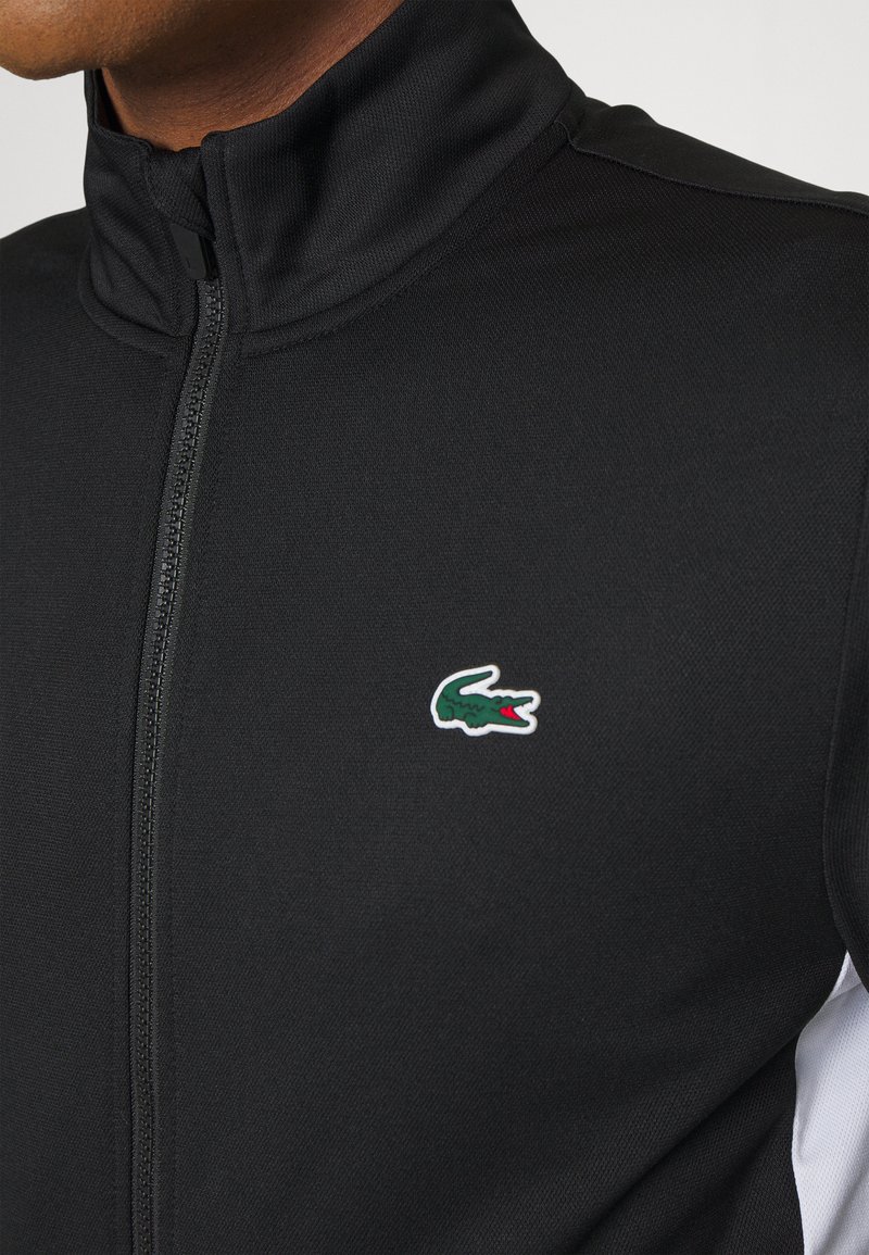 Zwarte zip-up jas met een hoge kraag, gemaakt van een gestructureerde stof en een groen krokodillenlogo op de borst; witte accenten aan de zijkanten.